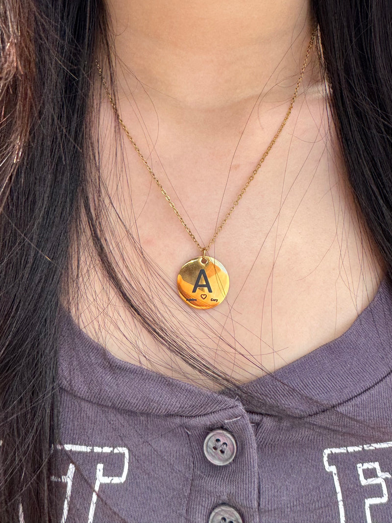 Person wearing a gold necklace with a round pendant featuring the letter 'A'.